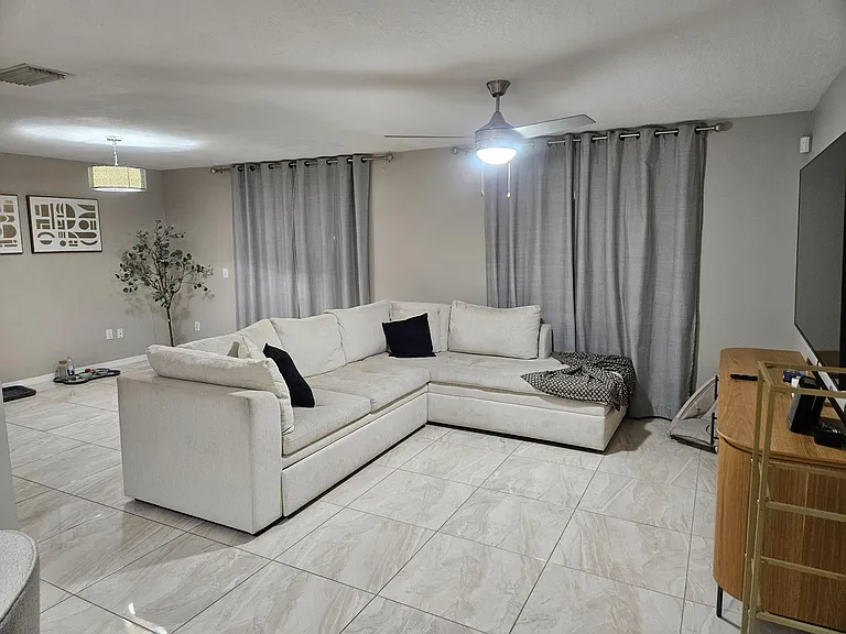 Spacious living area with a large sectional, tile flooring, ceiling fan, and wide window coverage with full-length curtains.