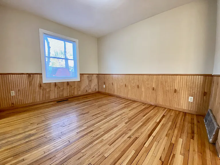 Bright room with polished hardwood floors and charming wood paneling, featuring a large window for abundant natural light. Bright room with polished hardwood floors and charming wood paneling, featuring a large window for abundant natural light.