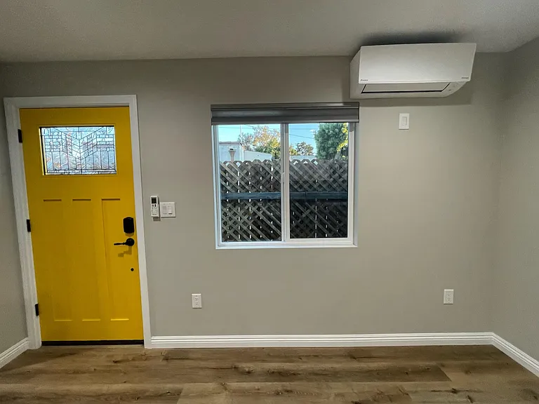 Living area with HVAC unit. Remote control near door for HVAC unit. Window looks onto outdoor space.