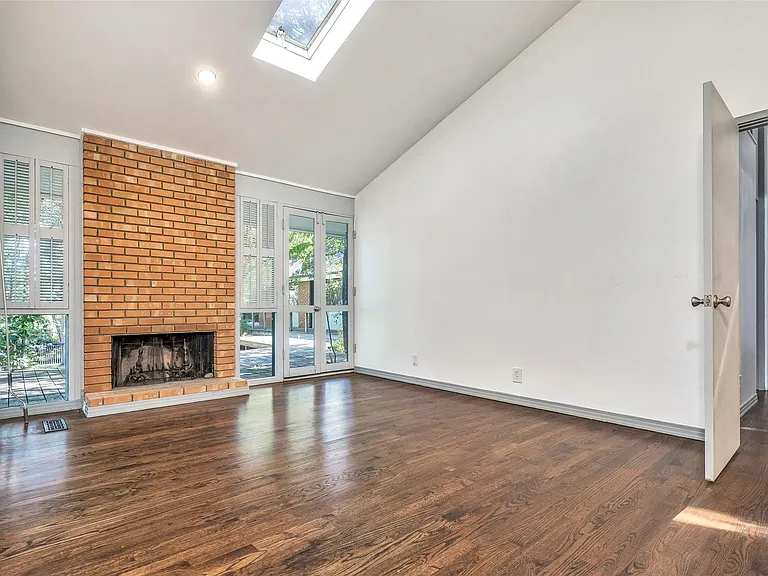 Spacious living area featuring a prominent brick fireplace with a raised hearth, wood-finish flooring, a vaulted ceiling, and a skylight