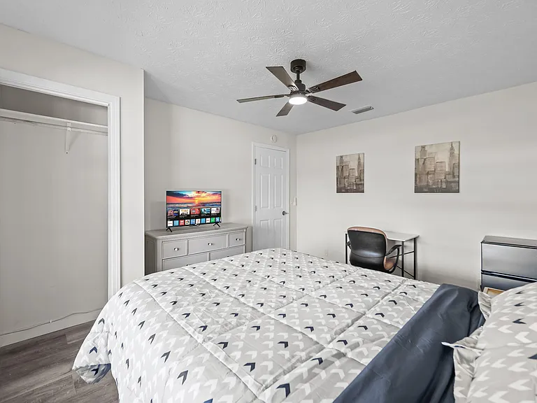 Furnished bedroom with a ceiling fan, spacious closet, dresser with TV, and a dedicated desk plus mini fridge.