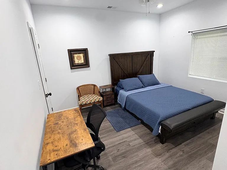 Furnished bedroom with ceiling fan, window blinds, and dedicated work desk plus nightstand and bench seating.