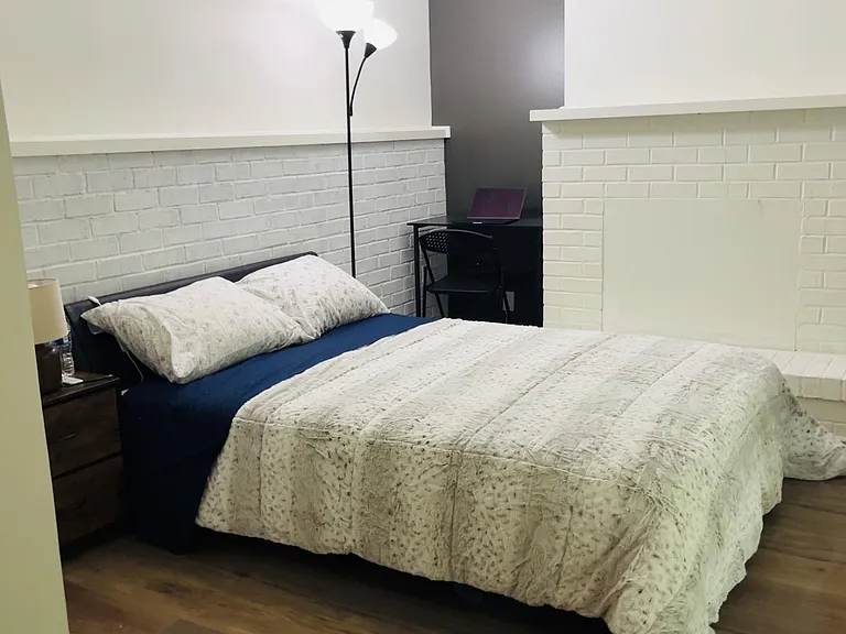 Furnished bedroom with queen bed, nightstand, and dedicated desk area, plus a painted brick fireplace feature wall.