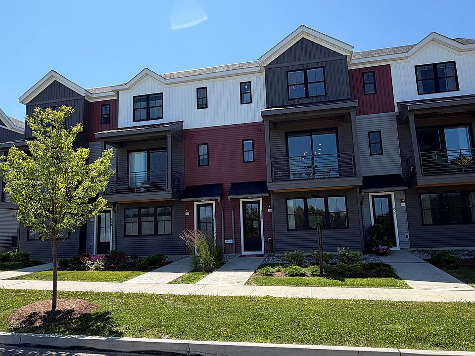 Modern three-story townhouse complex with balconies, a green lawn, and a young tree in front under a