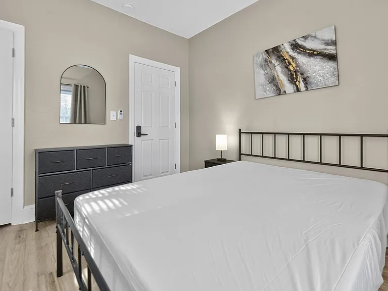 Furnished bedroom with a queen bed, nightstand lighting, dresser storage, and a wall mirror near the entry.
