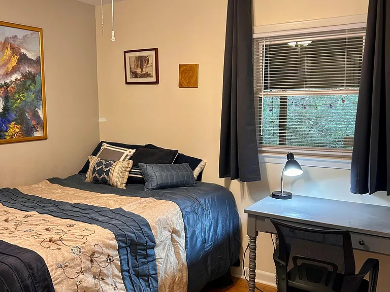 Well-appointed bedroom with a furnished workspace, hardwood floors, ceiling fan, and window with blinds and curtains.