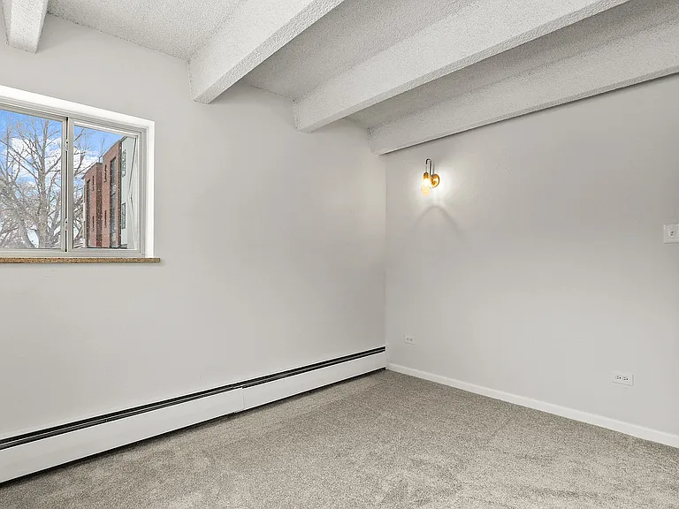 Bright and cozy room with large window, plush carpet, and modern sconce light, perfect for creating a personal retreat.