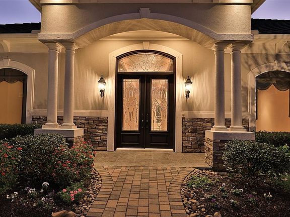 ELEGANT DOUBLE LEAD GLASS FRONT DOORS WITH ARCHED TRANSOM WINDOW - The front elevation of the home is beautiful with stucco exterior and stacked stone accents. The front door is gracefully framed by a stucco archway with double Doric columns.