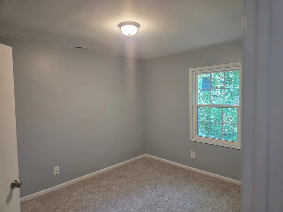 Bright and inviting bedroom featuring soft neutral carpeting, modern gray walls, and a large window that fills the space with natural light. The versatile layout provides ample room for a bed, dresser, and workspace, making it ideal as a guest room, child