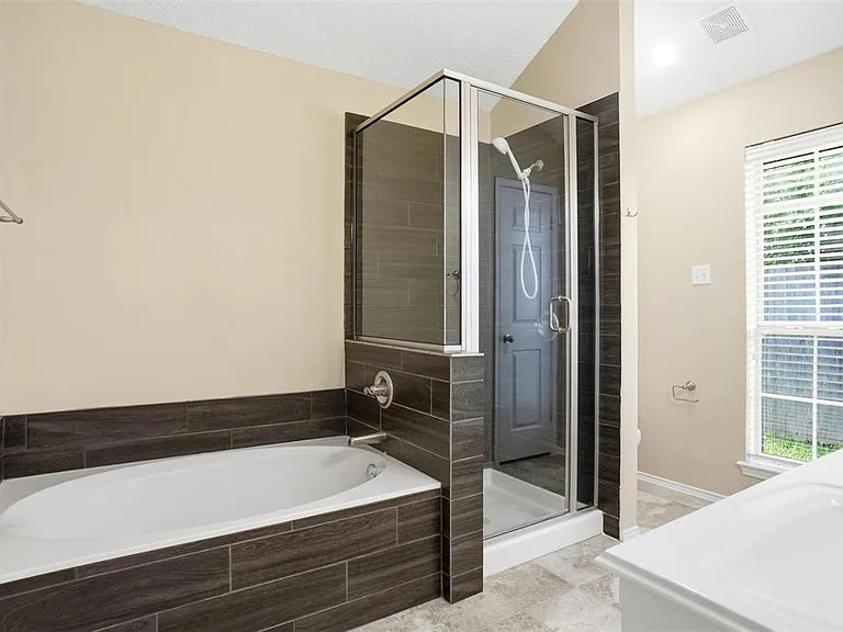 Bathroom featuring a built-in soaking tub with dark wood-look tile surround, a glass-enclosed shower with matching tile, and a window with blinds