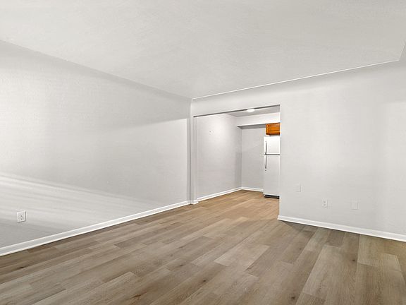 Bright and spacious open-plan living area with sleek laminate wood flooring and crisp white walls, leading to a cozy kitchen nook with modern appliances.