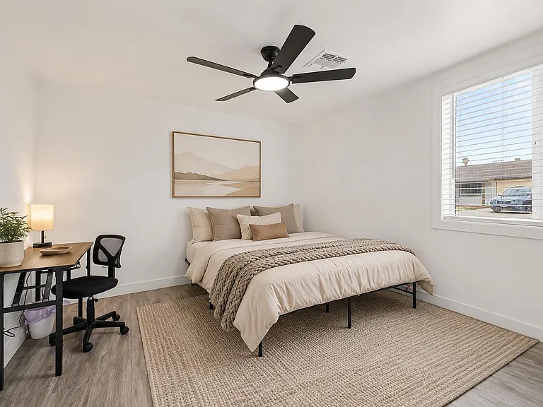 Furnished bedroom featuring a quee bed, dedicated desk workspace, ceiling fan, and a large window with blinds for natural lig