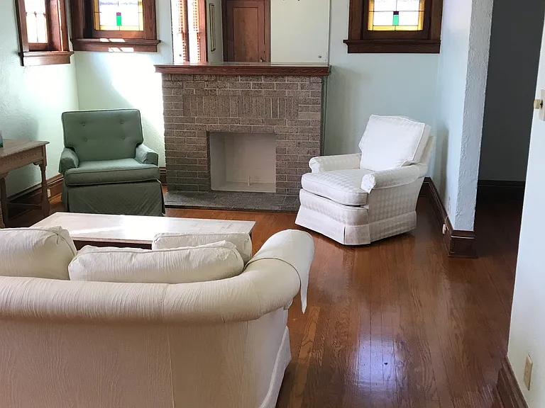 Living room, no furniture available. Ornamental fireplace, stained glass windows, ceiling fan. Wood Blinds. Southern exposure.