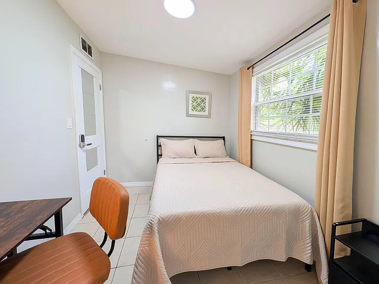 Furnished bedroom with a full-size bed, tile flooring, and a large window with blinds and curtains for natural light.
