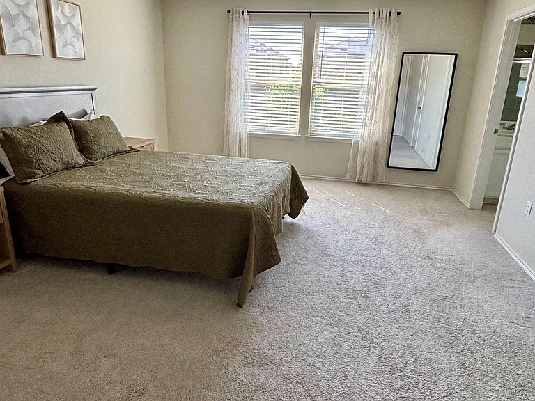 Spacious carpeted bedroom with a large window, ceiling light, and room for a full/queen setup plus extra furniture.