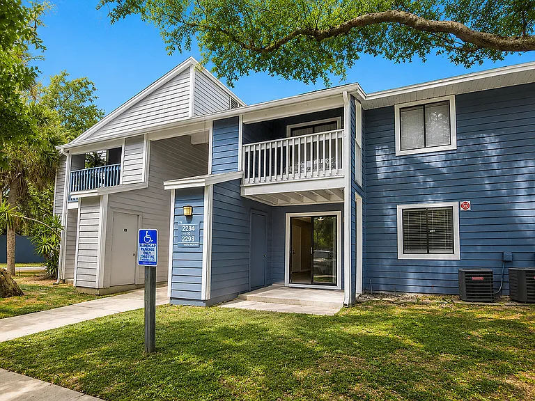 Charming blue apartment building with a welcoming porch and lush greenery, ideal for comfortable living.
