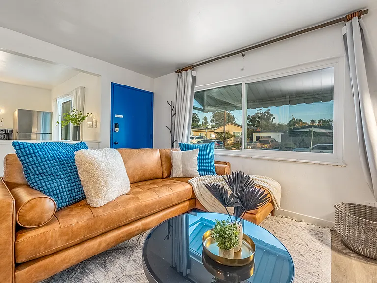 Large windows, quiet suburban street lined with houses and trees can be seen, adding to the homey and relaxed vibe of the main house living room.