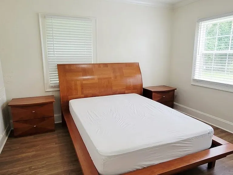 Spacious bedroom with a queen bed setup, matching nightstands, and multiple windows with blinds for natural light.