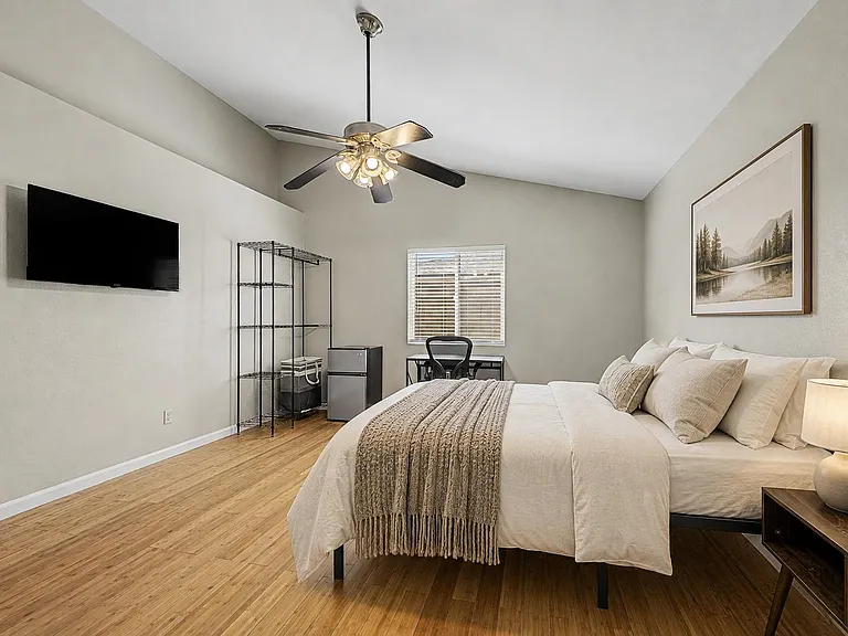 Spacious furnished bedroom with wood-style flooring, ceiling fan, desk area, mini fridge, and a wall-mounted TV.