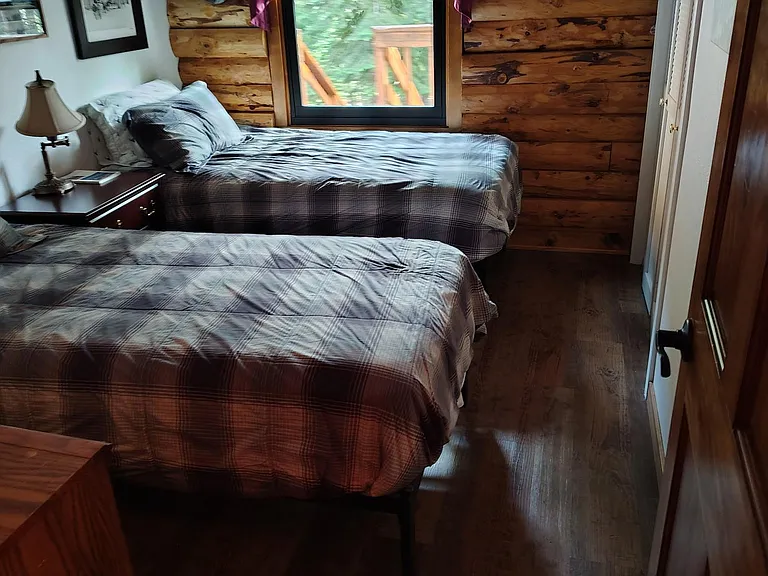 Bedroom with two twin beds, wood accent walls, and a window with wooded views; includes nightstand lighting and closet storag