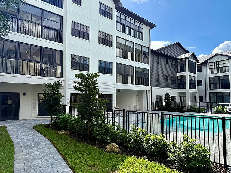 Modern apartment complex with large windows, lush greenery, and inviting pool under a clear blue sky.