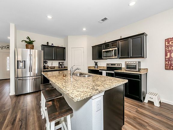 This modern kitchen features dark cabinetry, speckled granite countertops, and stainless steel appliances. A large central island with barstools anchors the space, which is finished with recessed lighting and warm wood-look flooring.