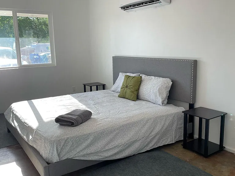 Furnished bedroom with a comfortable bed, nightstands, ceiling fan, and wall-mounted mini-split AC plus a large window.