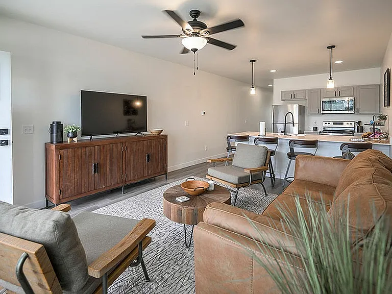 Chic and modern open-concept apartment living area featuring a cozy leather sofa, stylish armchairs, and a sleek wooden TV console with a flat-screen TV. The space flows into a contemporary kitchen with stainless steel appliances and elegant bar seating u