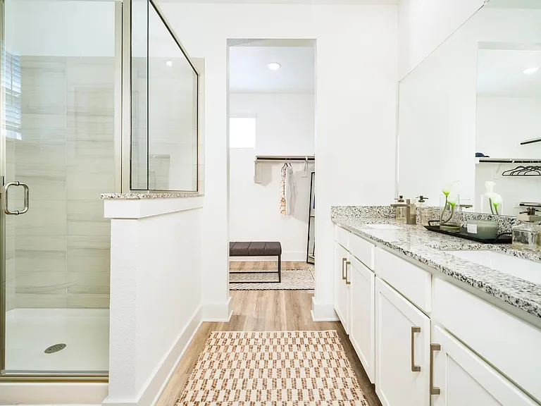 A bright primary bath features dual vanities, stone countertops, and a glass-enclosed shower, with a seamless connection to the spacious closet beyond.