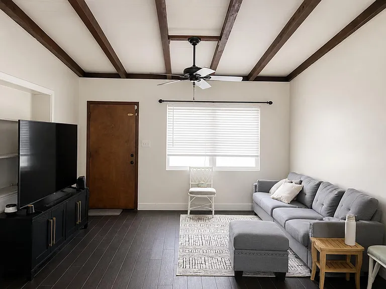 Common living area with exposed-beam ceiling, ceiling fan, and ample seating beside a large window with blinds.
