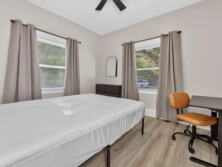 Furnished bedroom with a full bed, dresser, and dedicated desk area, plus two windows with blinds and curtains.
