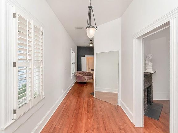 Hallway featuring wood-finish flooring, white plantation shutters, a suspended glass pendant light, and a framed doorway opening to a room with a dark stone fireplace