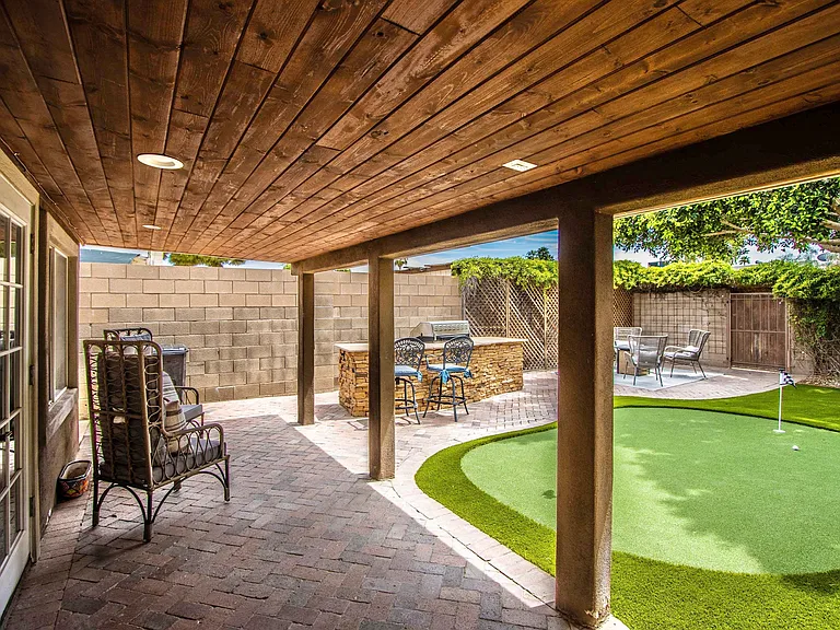 Shaded back patio at Casa Del Sol, Tempe rental home. Features a handsome wood-paneled ceiling with recessed lighting, brick pavers, high-back wicker chairs, and a direct view of the outdoor BBQ island. Perfect shaded escape for a Tempe, AZ stay.
