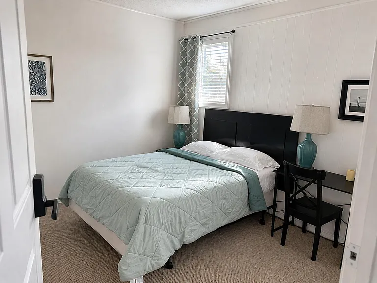 Furnished bedroom with a full-size bed, nightstands, and a small desk area, plus a window with blinds and curtains.