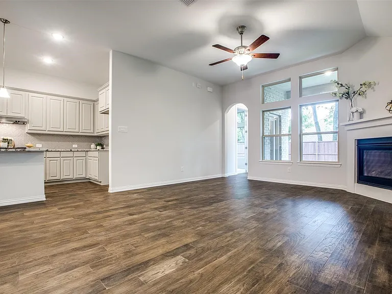 Open-concept living space featuring wood-finish flooring, a contemporary fireplace with a white mantel, and a kitchen with light-colored cabinetry and stone countertops
