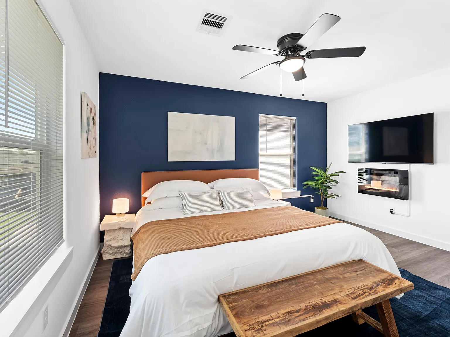 Spacious bedroom with large windows, ceiling fan, and a wall-mounted TV above an electric fireplace for added comfort.