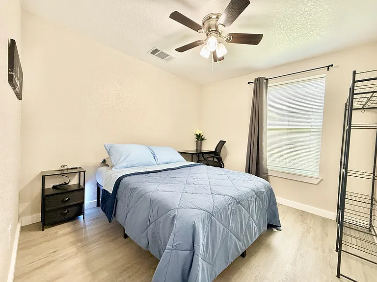 Furnished bedroom with a ceiling fan, large window with blinds, and a desk workspace for studying or remote work.