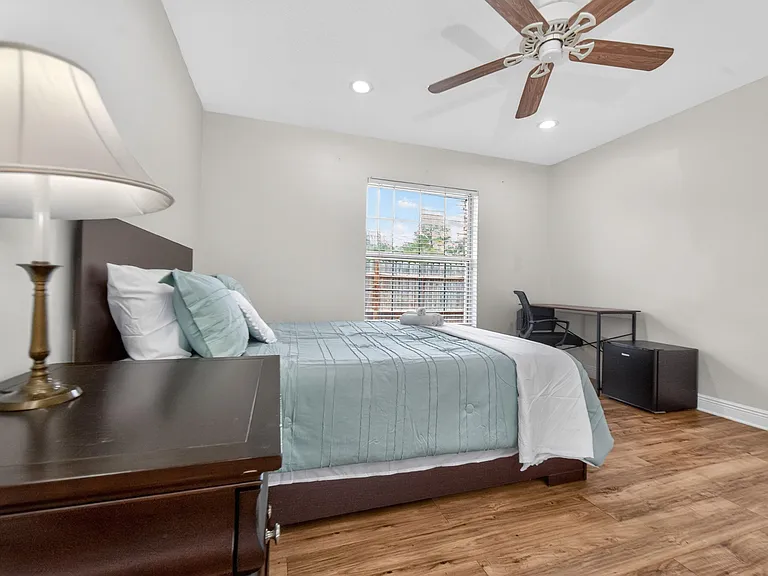 Well-appointed bedroom with a ceiling fan, hardwood-style floors, and a large window; includes a work desk and mini fridge.