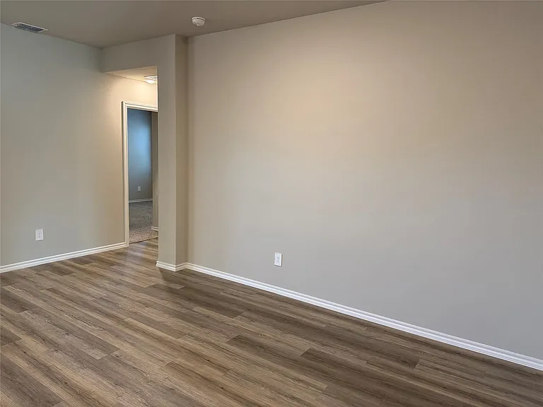 Spacious room featuring wood-finish flooring, light neutral wall paint, white baseboards, and a doorway leading to a carpeted room