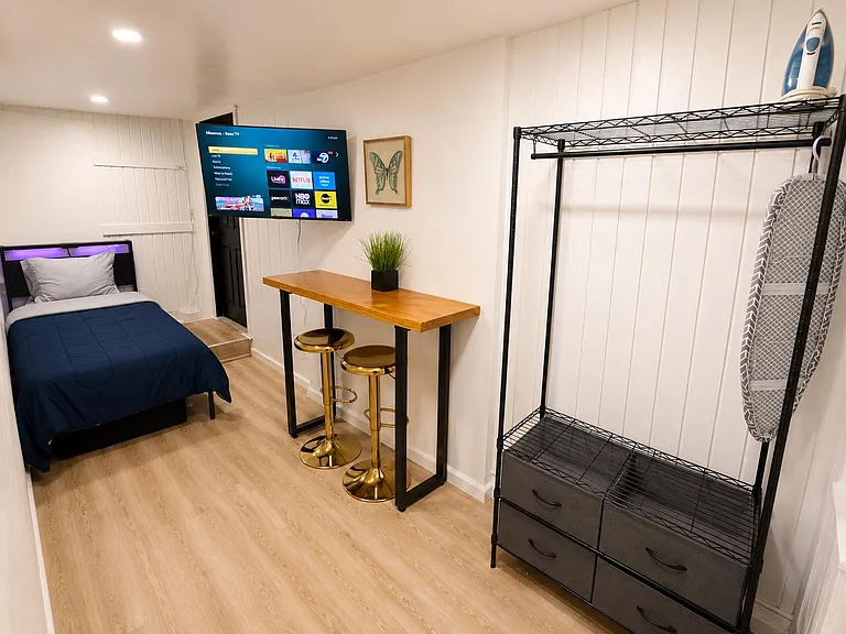 Furnished bedroom with twin bed, wall-mounted TV, bistro desk with stools, and open closet storage with drawers.