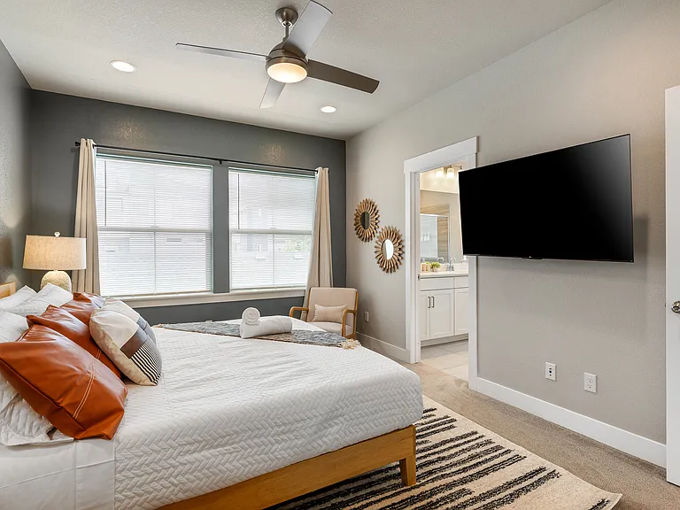 Bright and spacious bedroom with a comfortable wooden bed, layered with white bedding and accent pillows. Enjoy natural light from large windows, a cozy armchair, a ceiling fan, and a wall-mounted TV for your relaxation and entertainment.