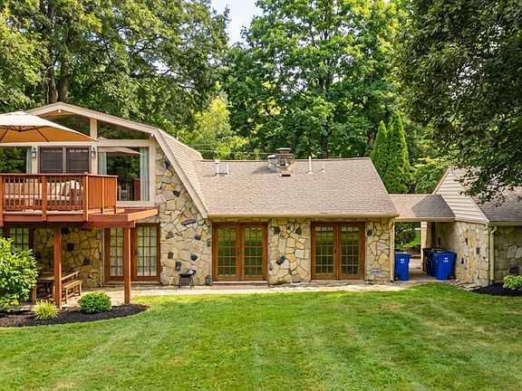 Rear view of the home featuring a beautiful stone exterior, expansive lawn, and a large elevated deck with covered seating. Surrounded by mature trees, this space offers privacy and a perfect setting for outdoor entertaining. 1.1acre site