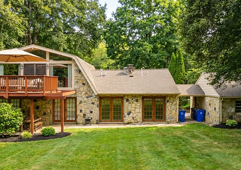 Rear view of the home featuring a beautiful stone exterior, expansive lawn, and a large elevated deck with covered seating. Surrounded by mature trees, this space offers privacy and a perfect setting for outdoor entertaining. 1.1acre site