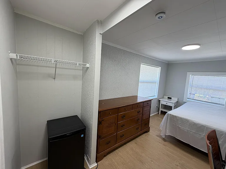 Furnished bedroom with a bed, dresser, nightstand, and a closet area with shelving plus a mini fridge for added convenience.