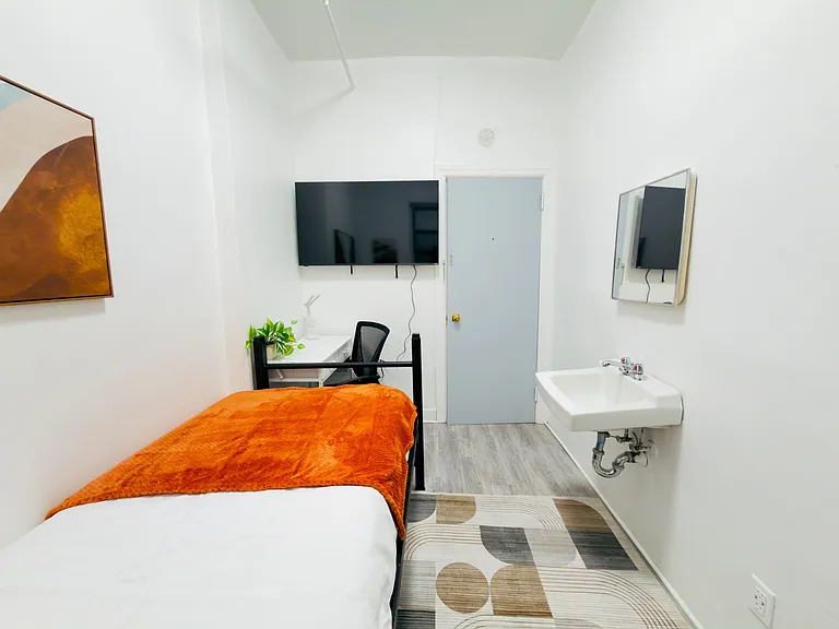 Private furnished bedroom with a wall-mounted TV, compact work desk, and convenient in-room sink plus mirrored storage.