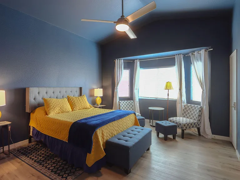 Spacious bedroom with a queen bed, ceiling fan, and seating area by large windows with curtains and blinds.