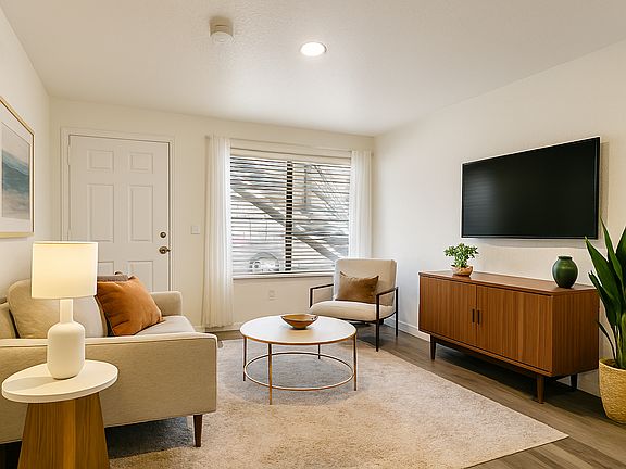Bright, modern living space with clean lines, natural light, and warm wood flooring. Features a large window with blinds and sheer curtains, recessed lighting, and a spacious layout ideal for flexible furniture arrangements and personal styling.
