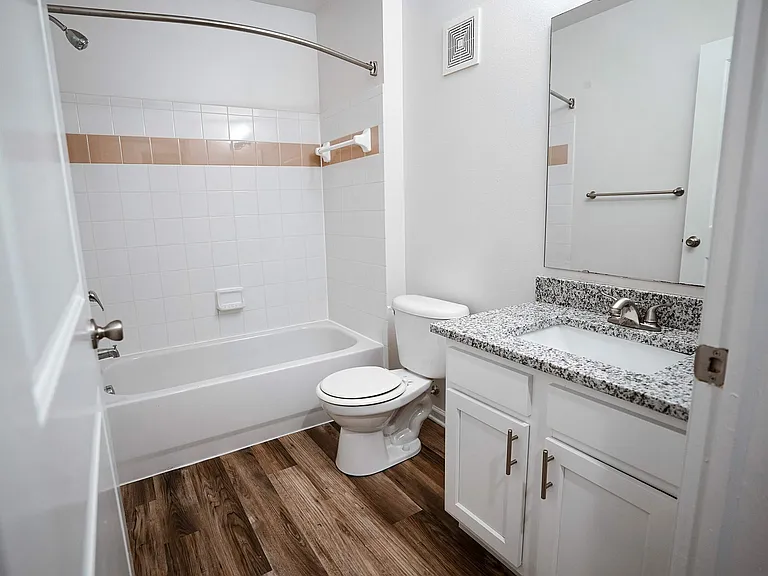 Enjoy the clean, bright feel of this bathroom at The Bristol _ featuring a granite countertop sink, hardwood-style flooring, and a shower/tub with crisp white cabinetry and fixtures.