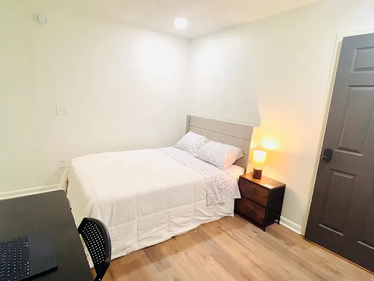 Furnished bedroom featuring a full bed, nightstand with lamp, and a dedicated desk workspace on easy-care flooring.