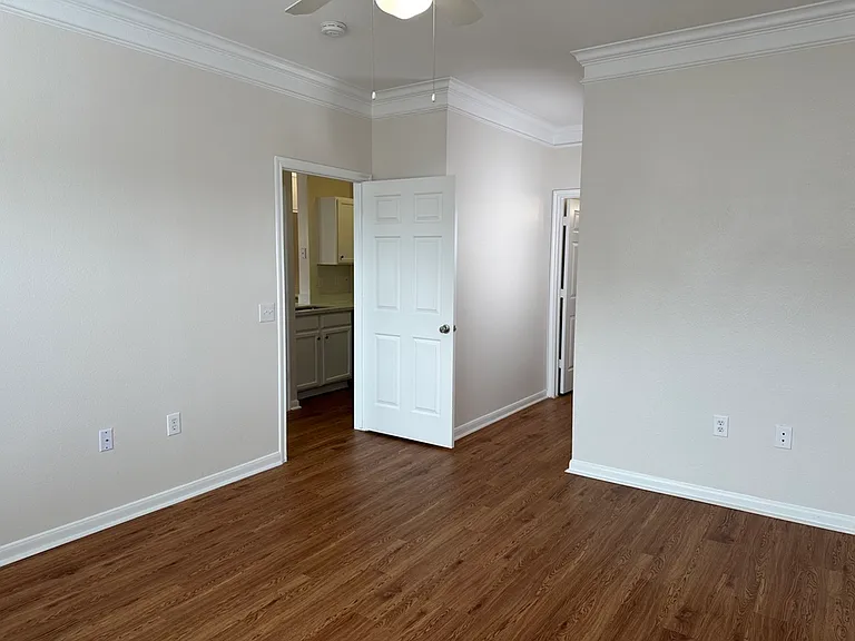 Bright and spacious room featuring elegant crown molding, wood-style floors, and an inviting white interior door leading to a modern kitchen area. Bright and spacious room featuring elegant crown molding, wood-style floors, and an inviting white interior 